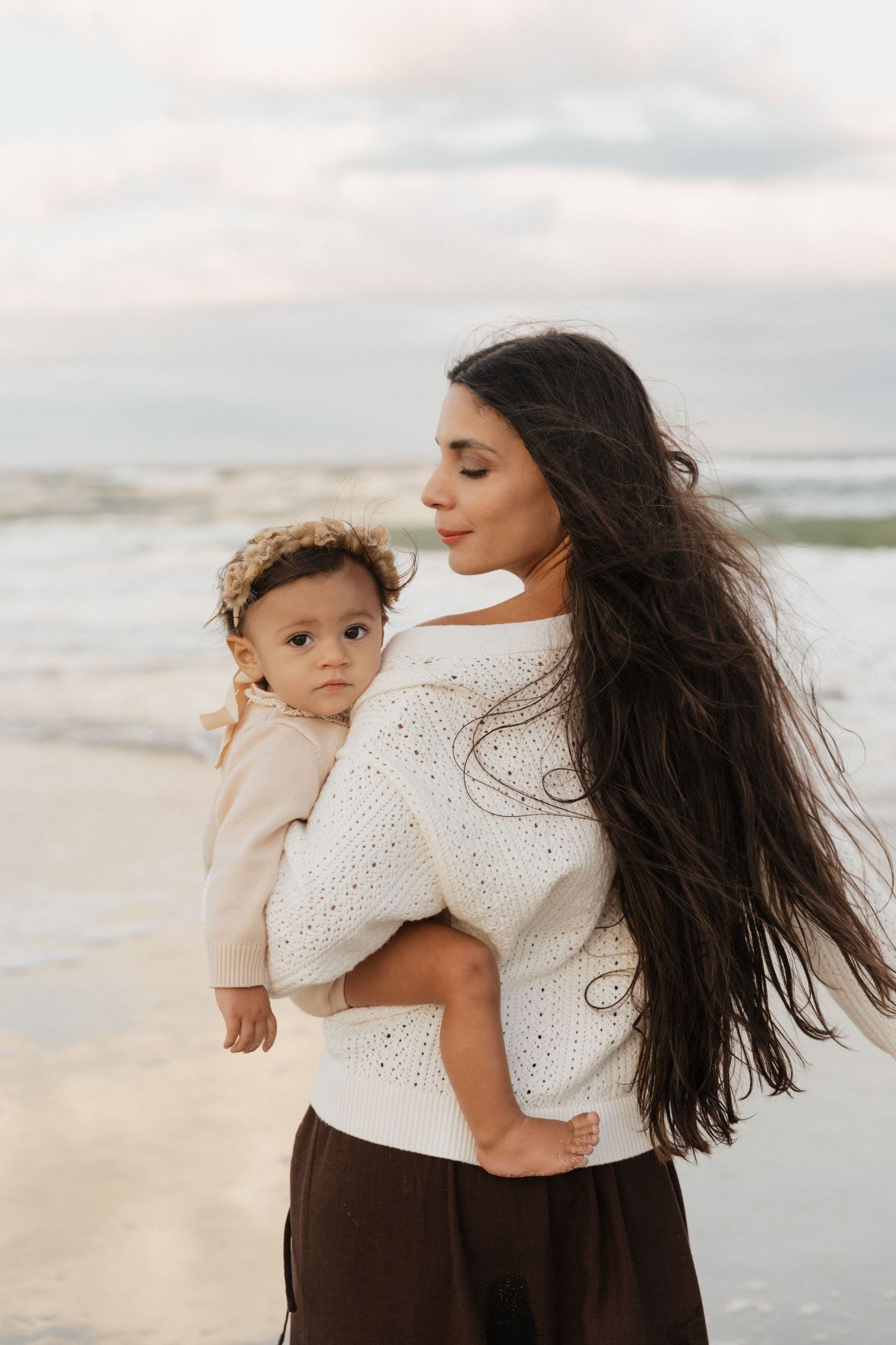 Mother and baby in matching white outfits share intimate moments against a beach backdrop with flowing hair.