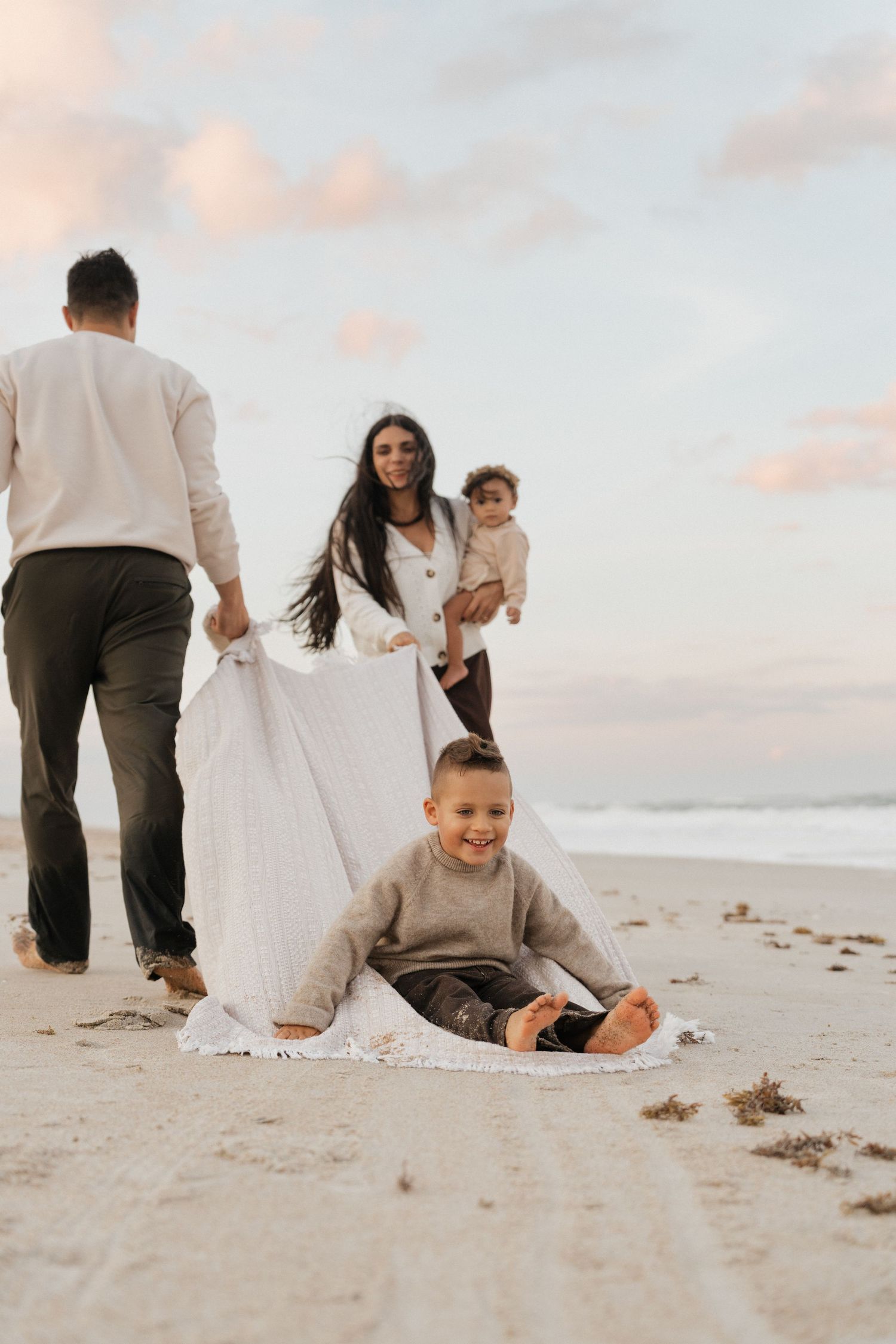 Candid family moments captured during sunset on a sandy beach with soft pink skies.