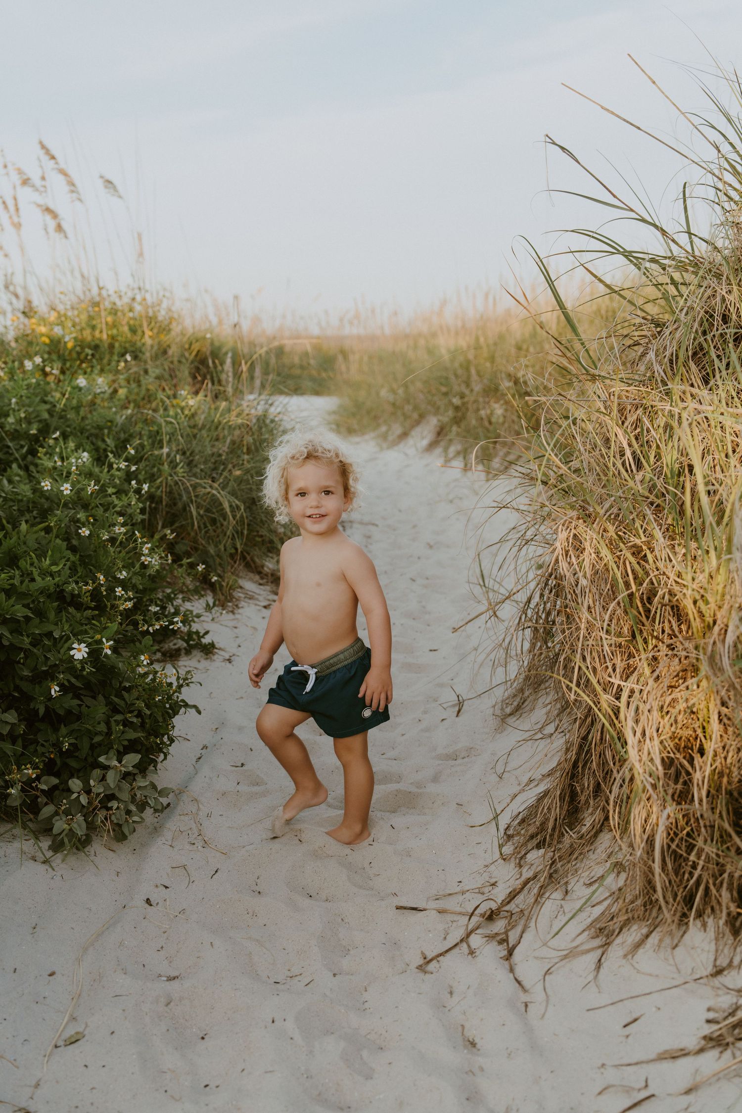 A small child walks barefoot on a sandy beach path surrounded by tall beach grass and dunes at sunset.