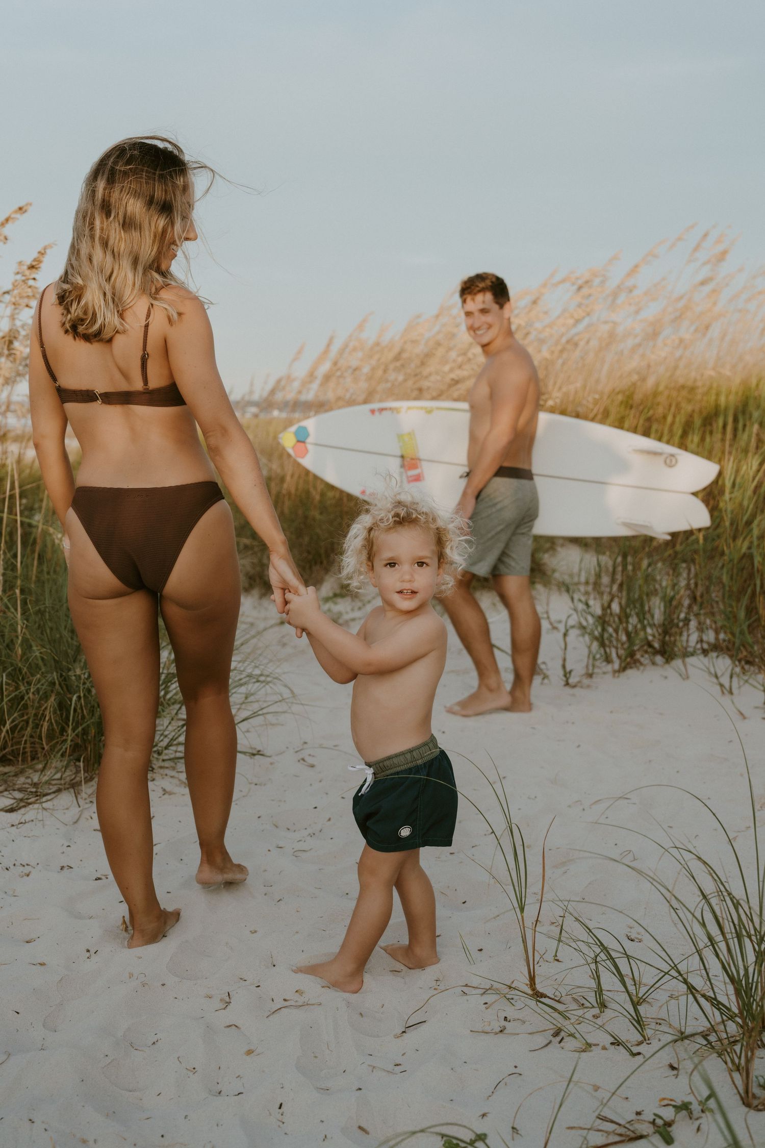 Family enjoying a sunny day at the beach with surfboards and tall grass on sandy dunes.