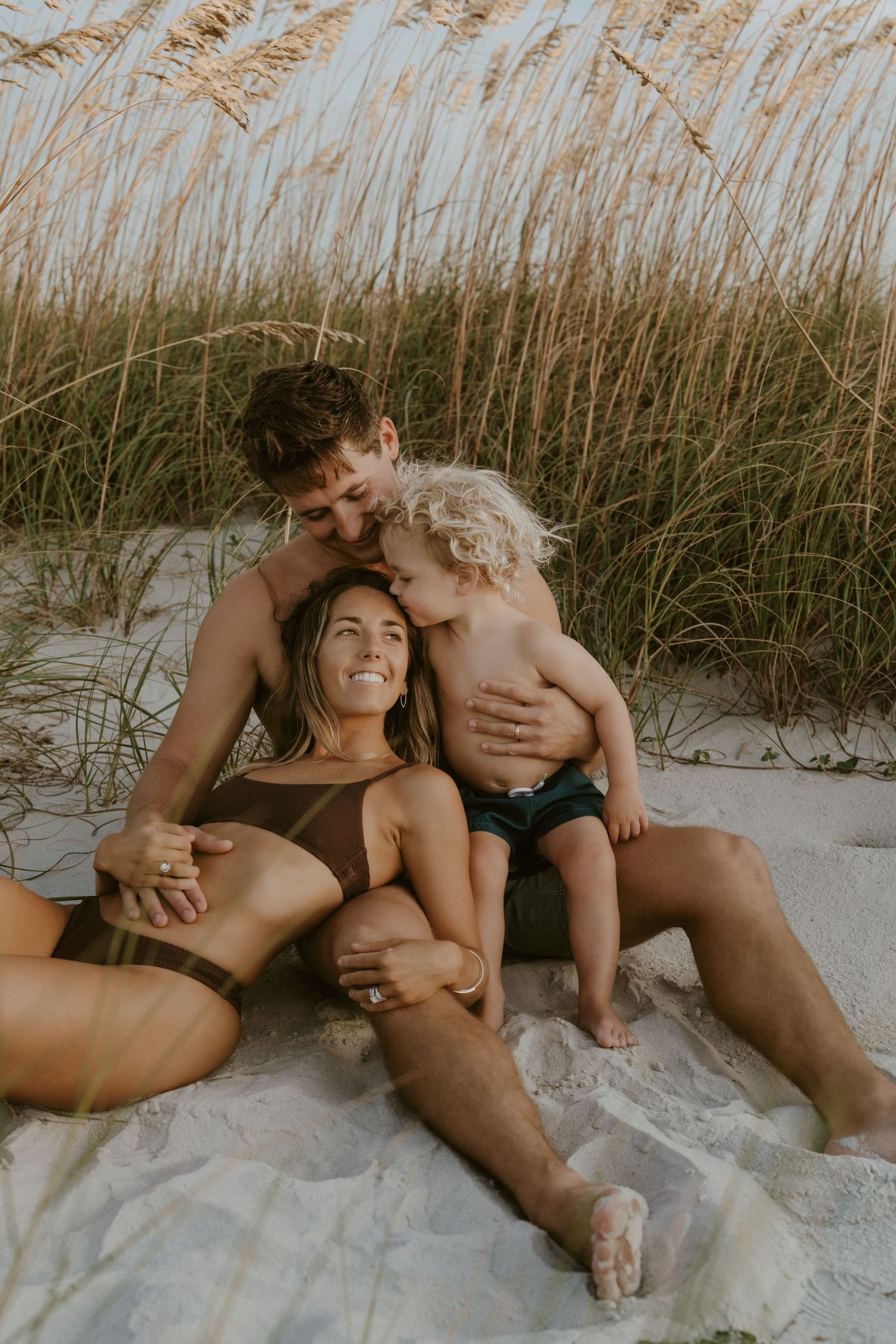 Intimate family moment captured on a sandy beach with sea grass swaying in the background.