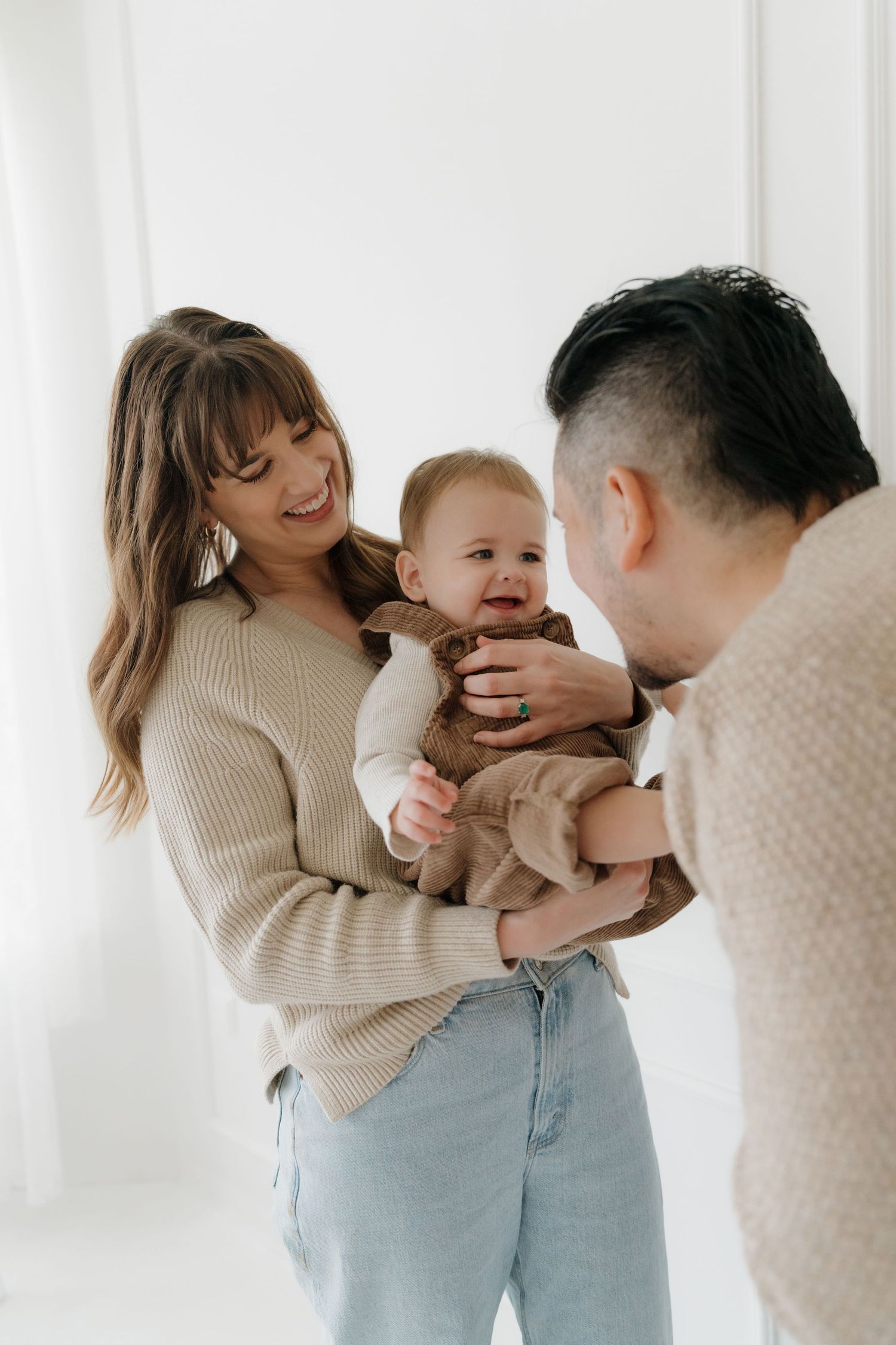 A family shares tender moments in matching beige sweaters and light denim against a bright white background.