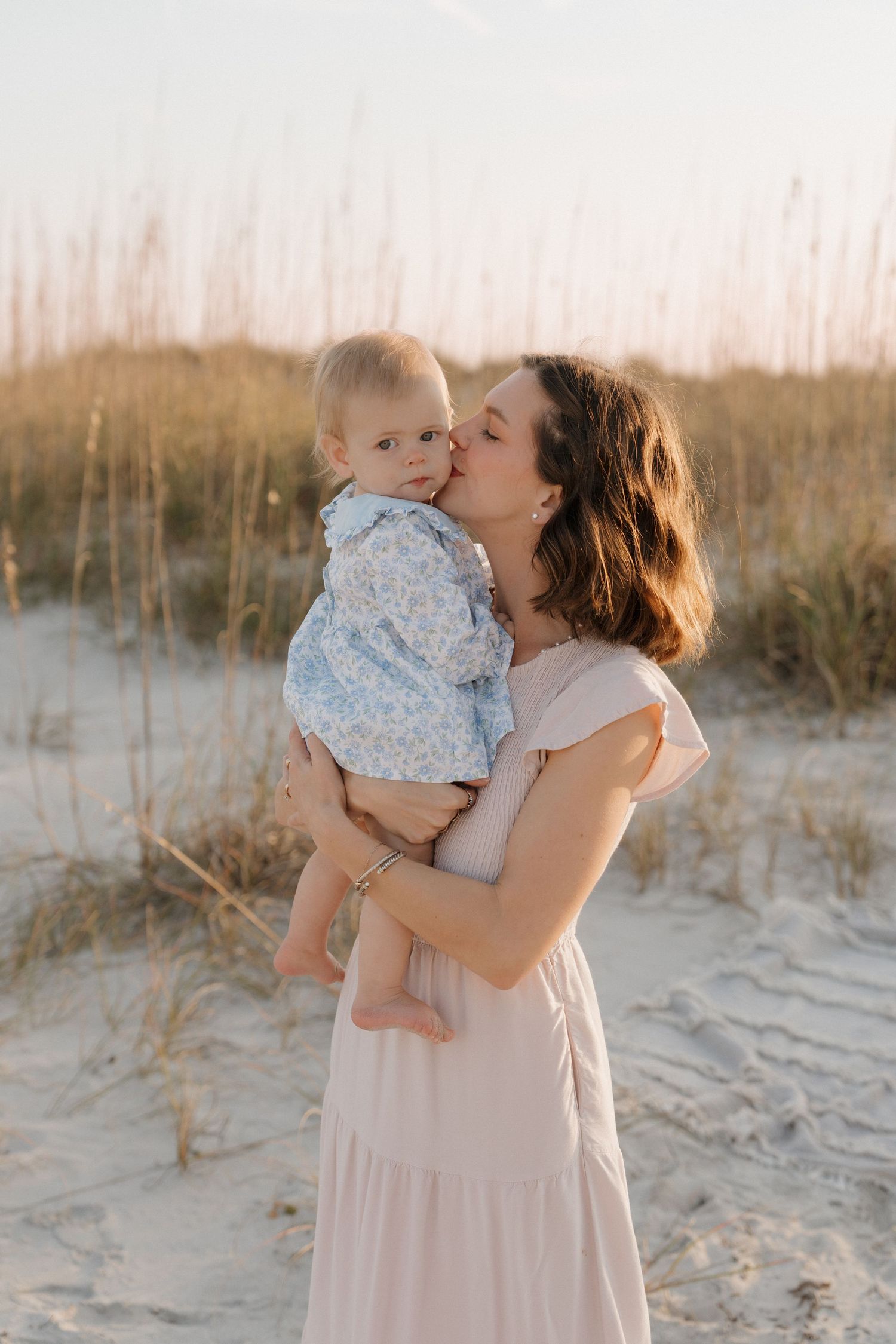 A tender beach scene with a parent and baby in light flowy clothing against sand dunes at sunset.