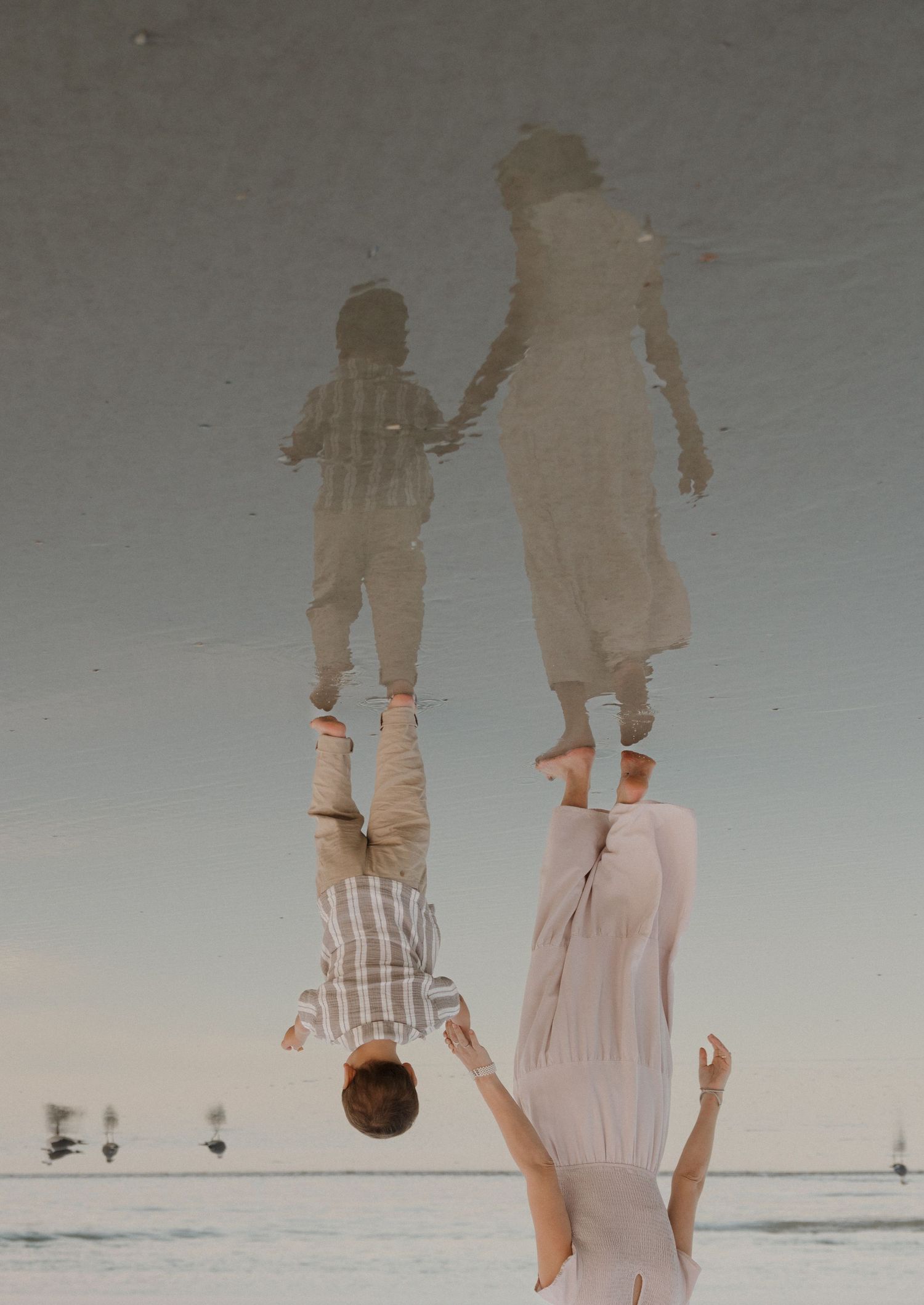 Two people in light colored clothing walking on a reflective wet beach surface at sunset.