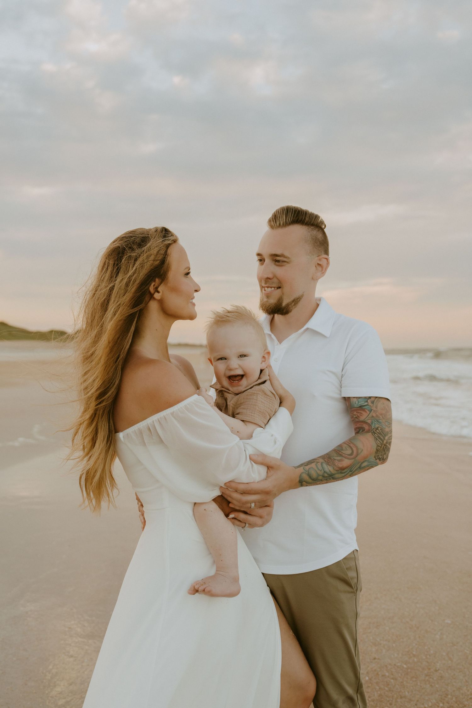 A couple shares a tender moment with their baby on a serene beach during golden hour.