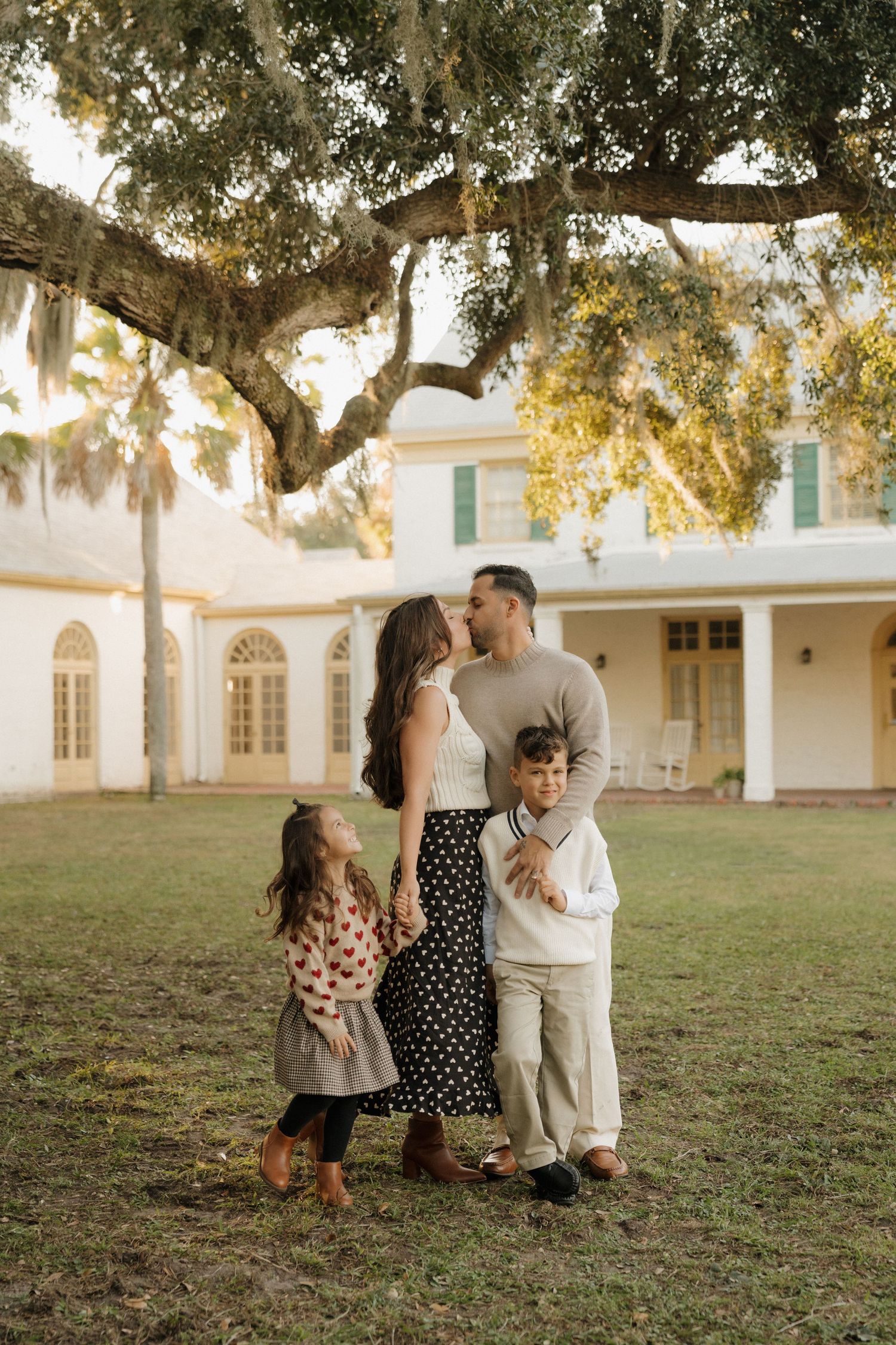 Family photoshoot under golden oak trees with white colonial style building in background.