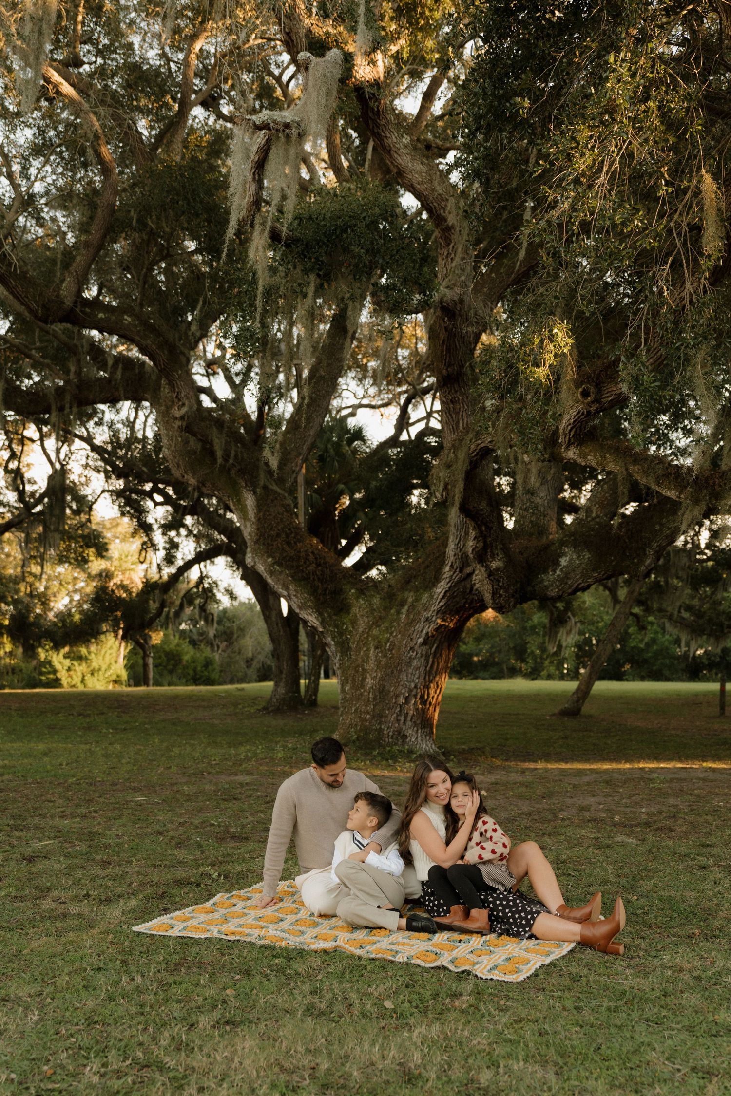 Family enjoying a relaxing picnic on a blanket under large oak trees in a grassy park.