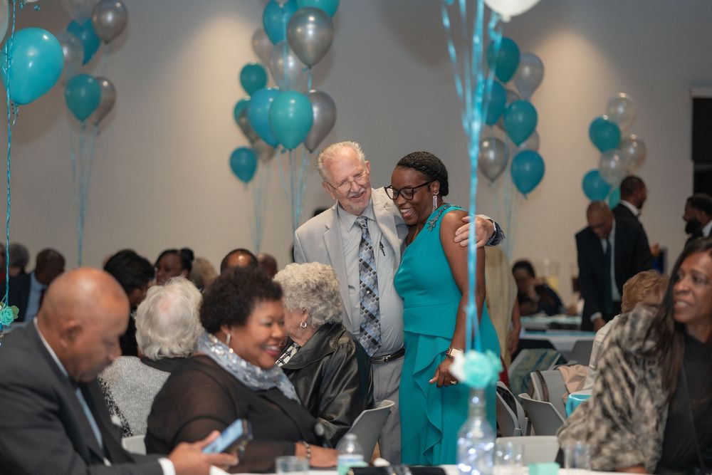 People gather at a celebration event decorated with teal balloons in a banquet setting.