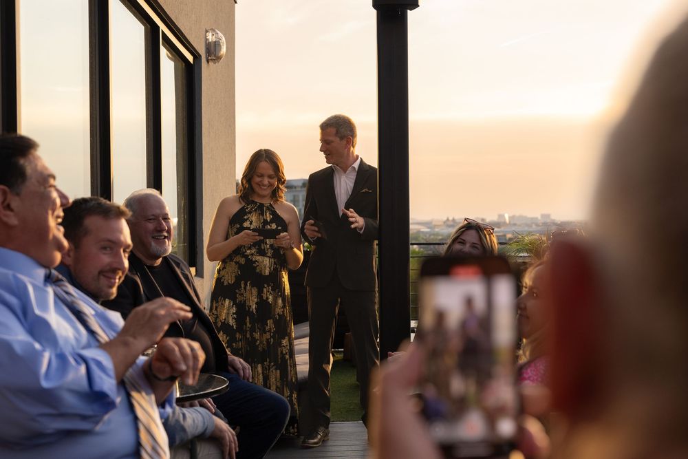 Couple speaks to guests at an outdoor rooftop party during sunset, with attendees photographing the moment.