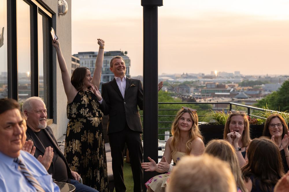 Celebratory rooftop gathering at sunset with guests applauding a couple raising their arms in joy.