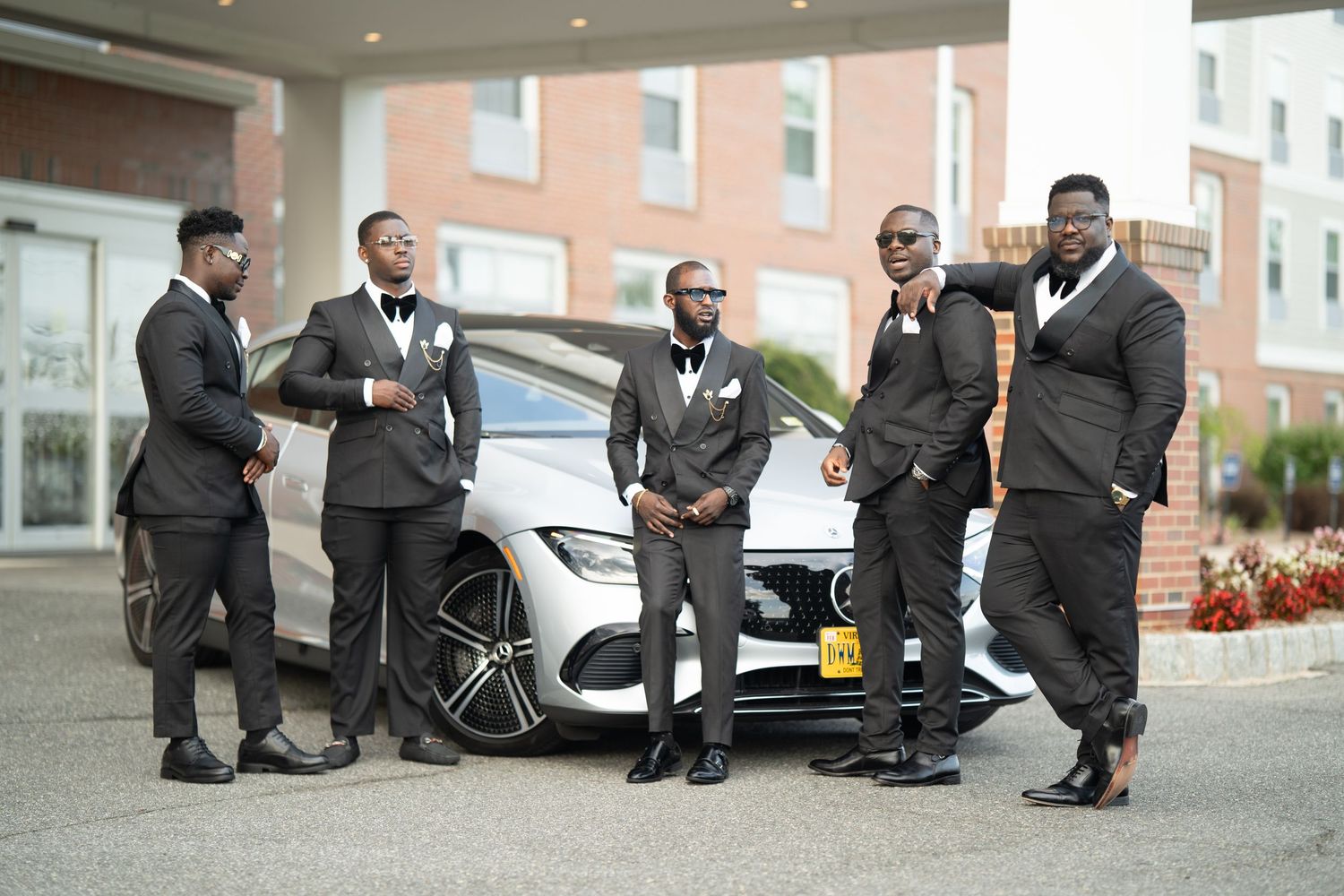 A group of men in formal black suits pose together in front of a luxury car.