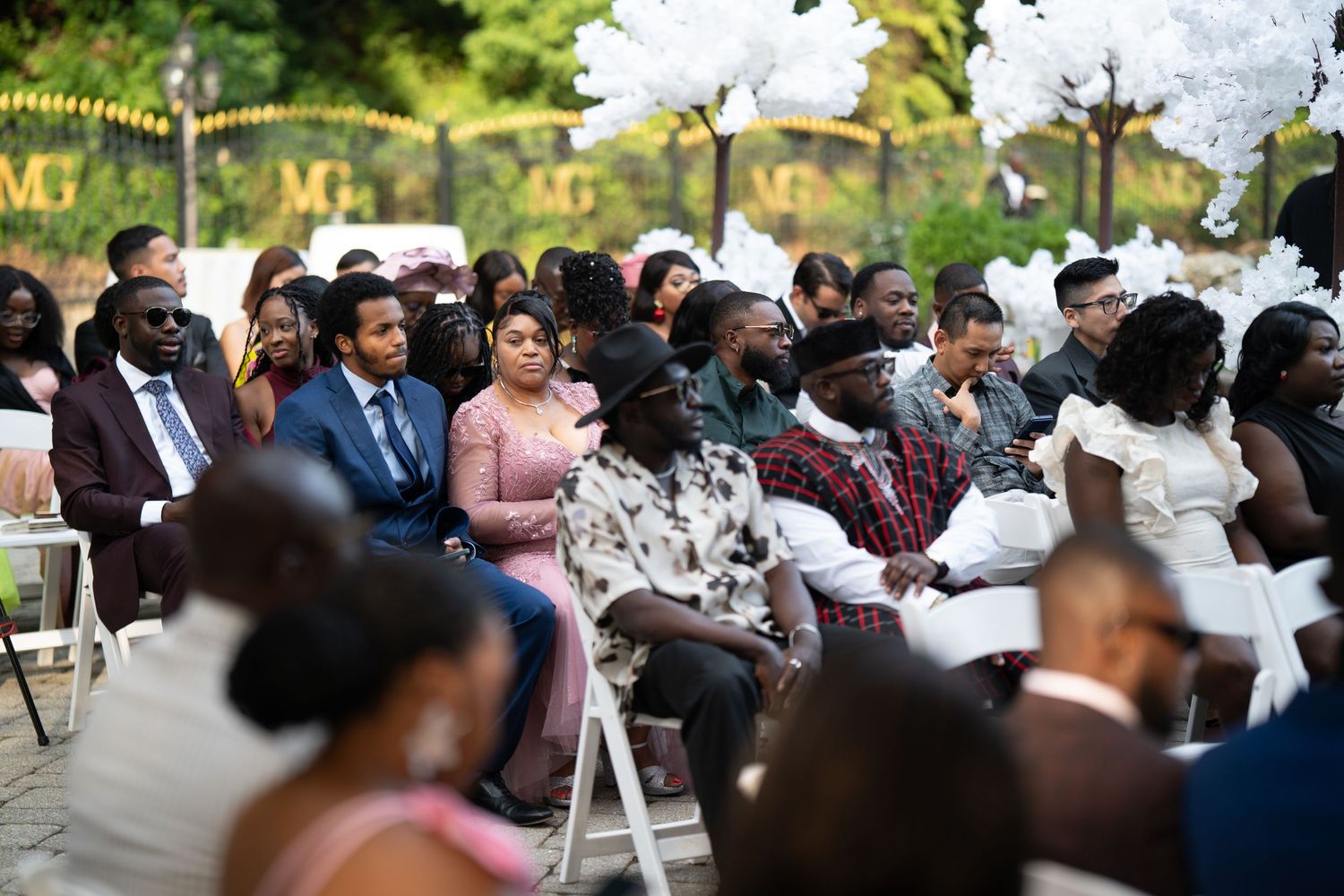 Wedding guests seated in white chairs outdoors with decorative white feather plumes visible during outdoor ceremony.