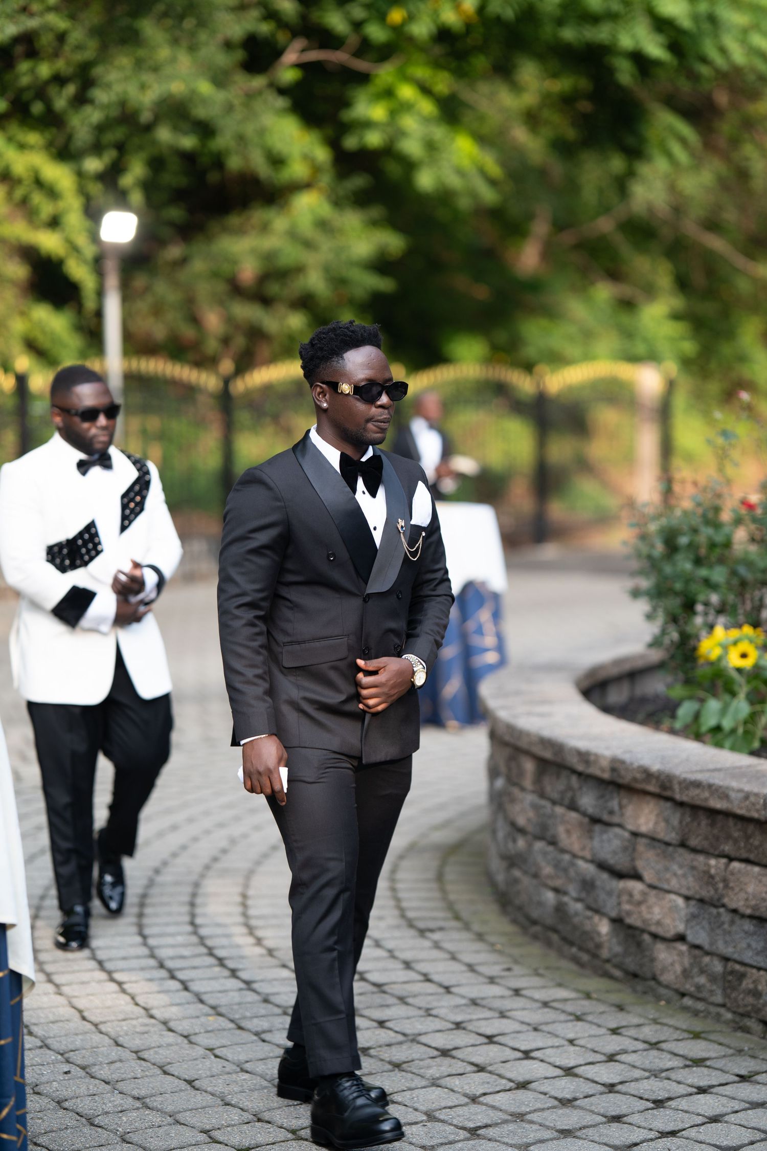 Groomsmen in black and white formal attire walk along a curved stone pathway in a garden setting.