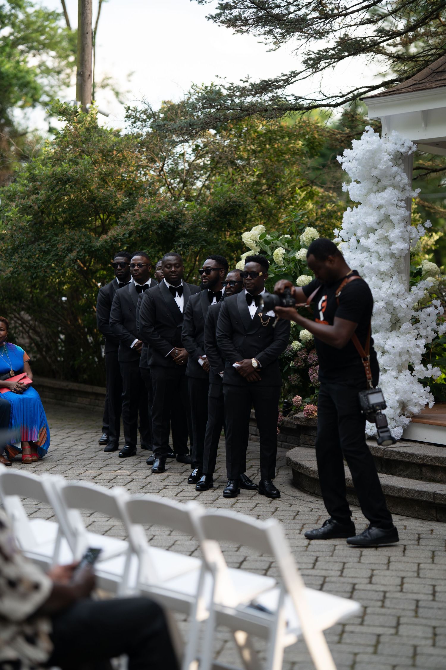 A wedding party in formal black attire stands in line on a stone patio with white balloon decorations.