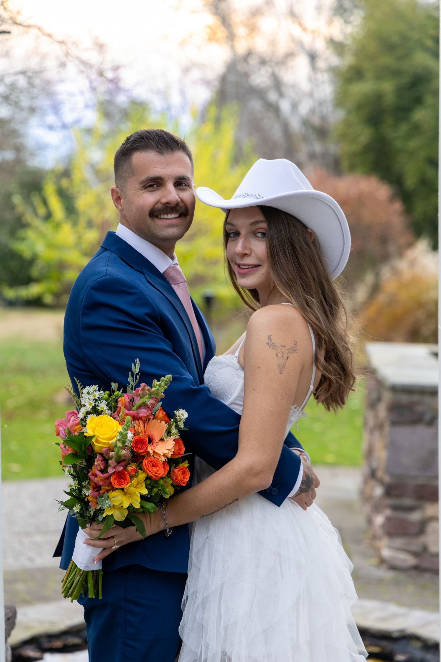A couple poses together outdoors in wedding attire with a bright bouquet of yellow and orange flowers.