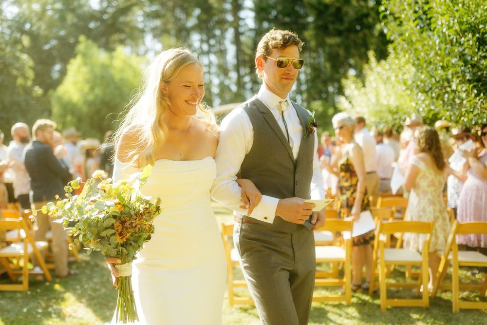 Outdoor wedding ceremony in a sunlit garden setting with guests seated on wooden chairs.