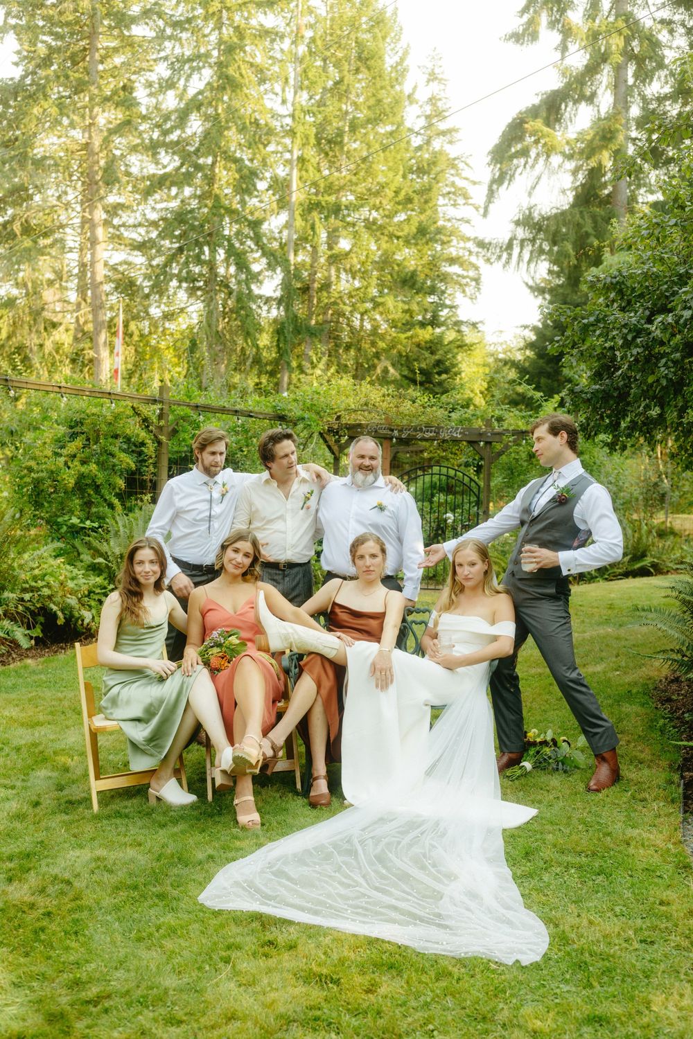 Wedding party relaxes on wooden benches in a garden setting during golden hour.