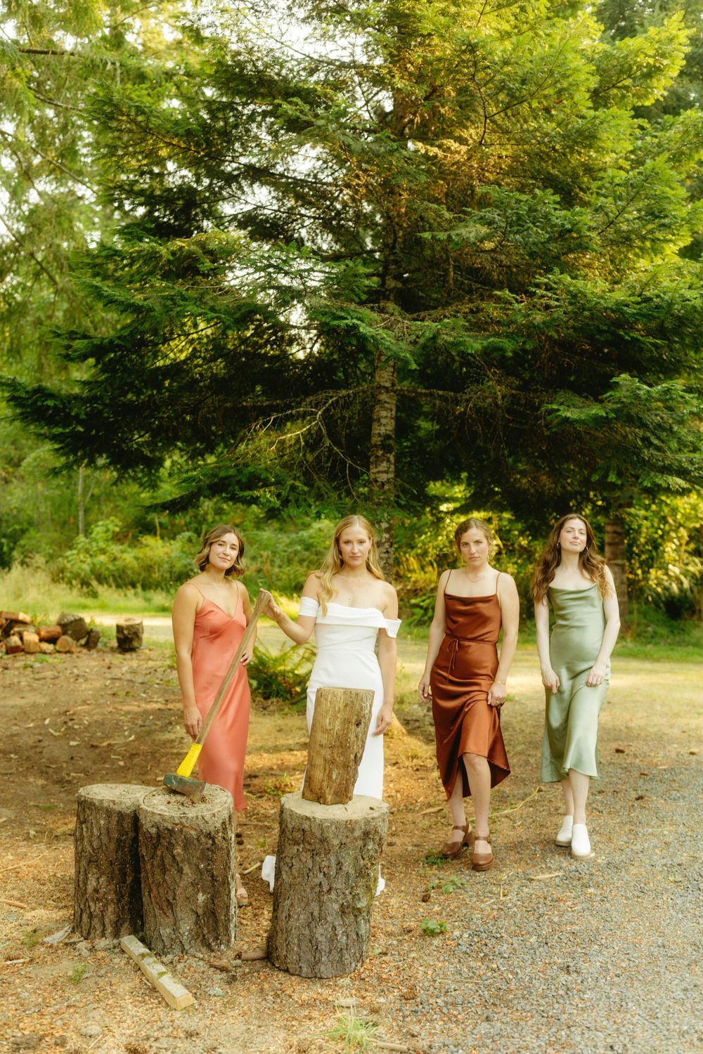 Four people in colorful dresses stand on tree stumps in a forest clearing.