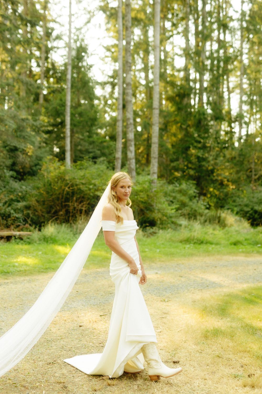 A person in a flowing white wedding dress walks along a forest path with a long trailing veil.