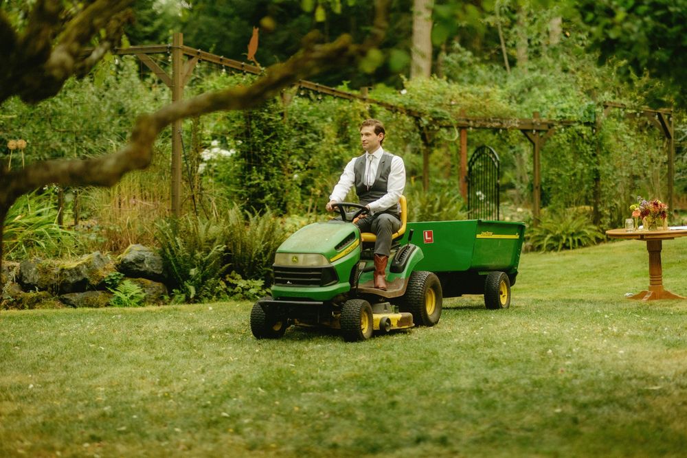 A person riding a green lawn tractor through a lush garden landscape.