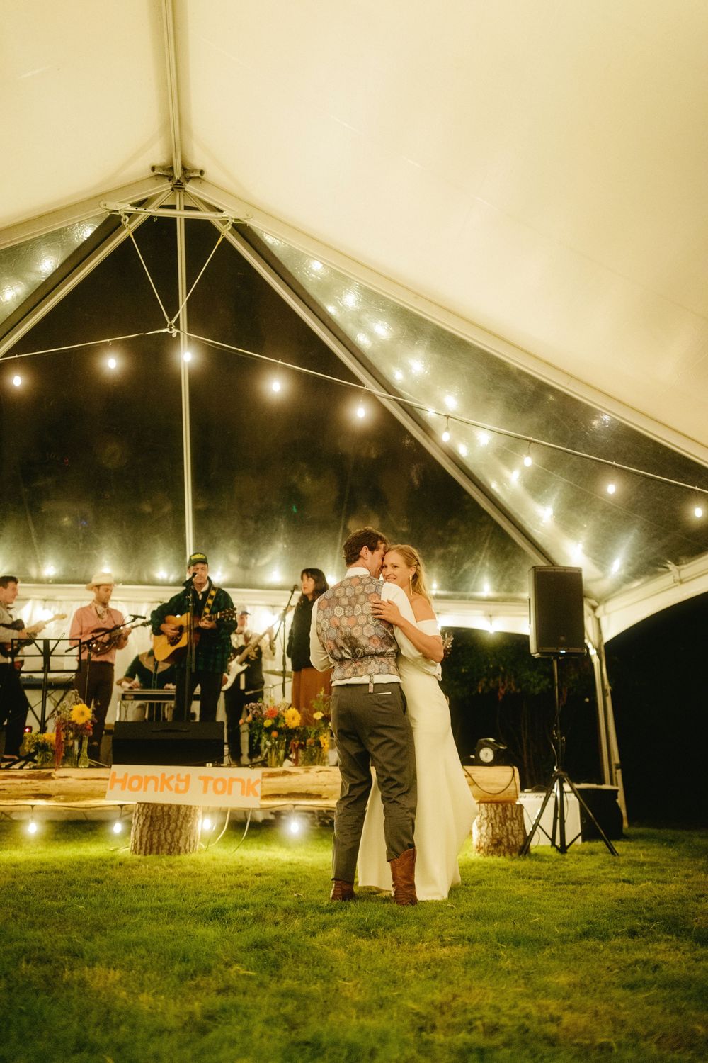 A couple dancing under string lights at an outdoor tent reception with a band performing in the background.