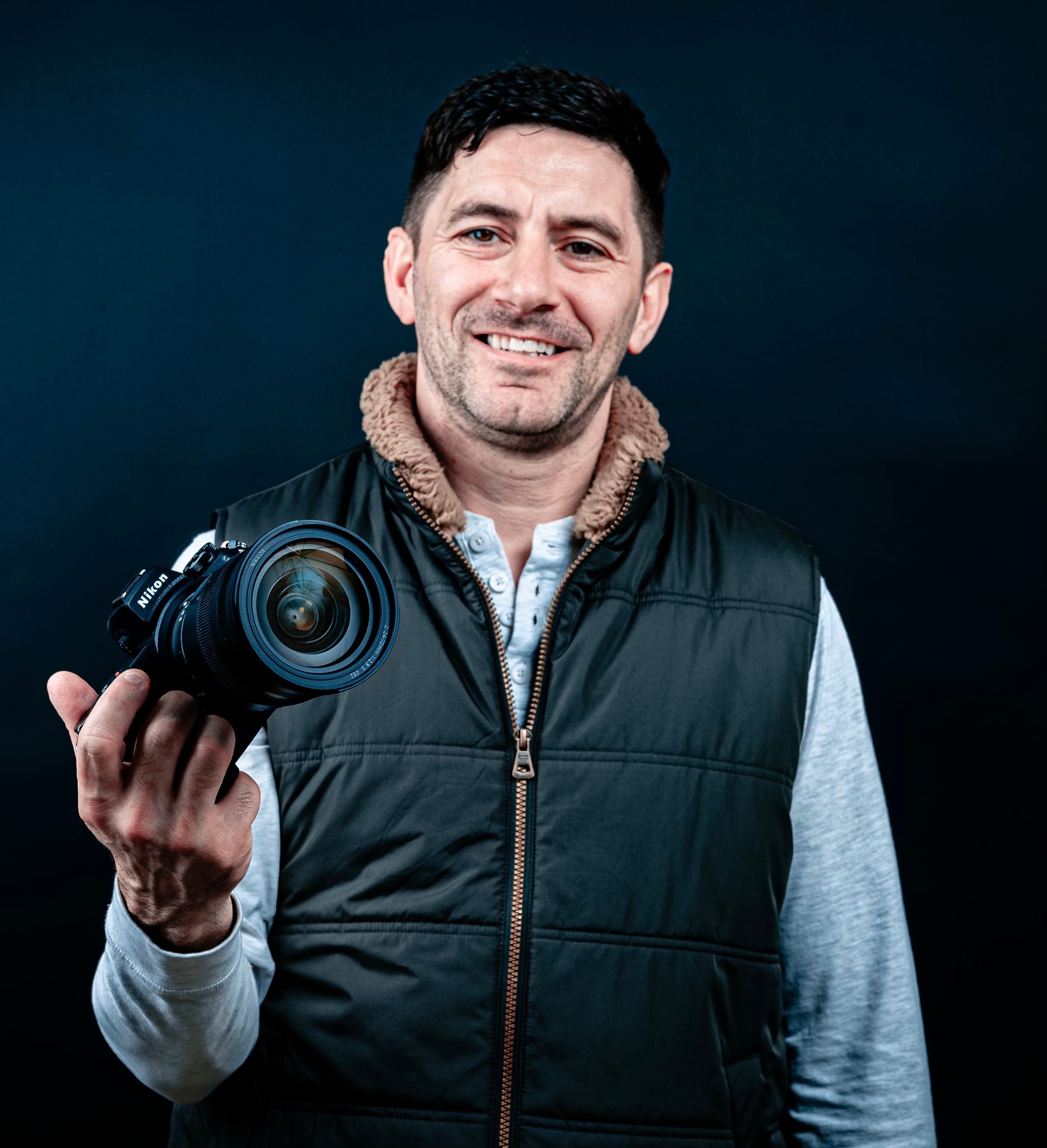 Person wearing a blue sweater smiles warmly while posing outdoors in natural lighting.