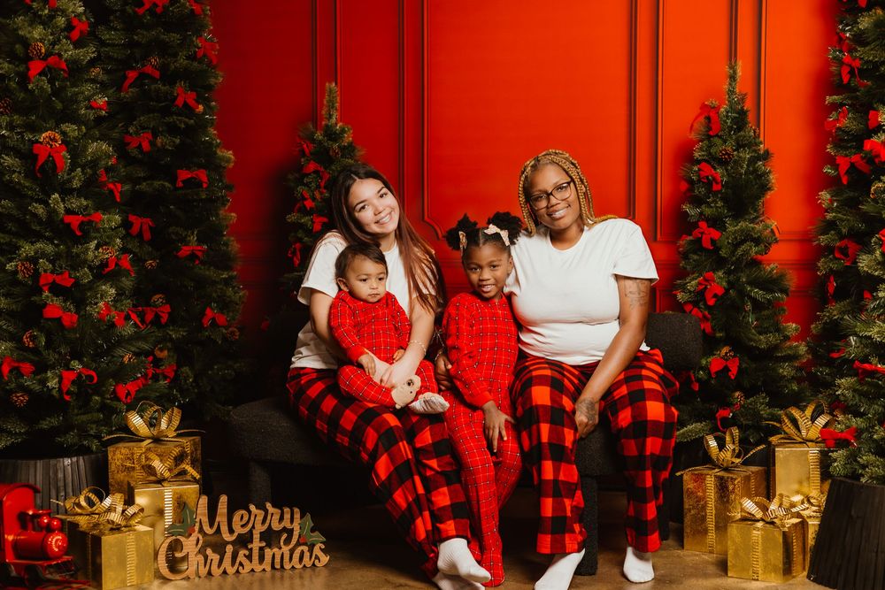 A festive Christmas photo session with a red backdrop, decorated tree, plaid blanket, and golden wrapped presents.