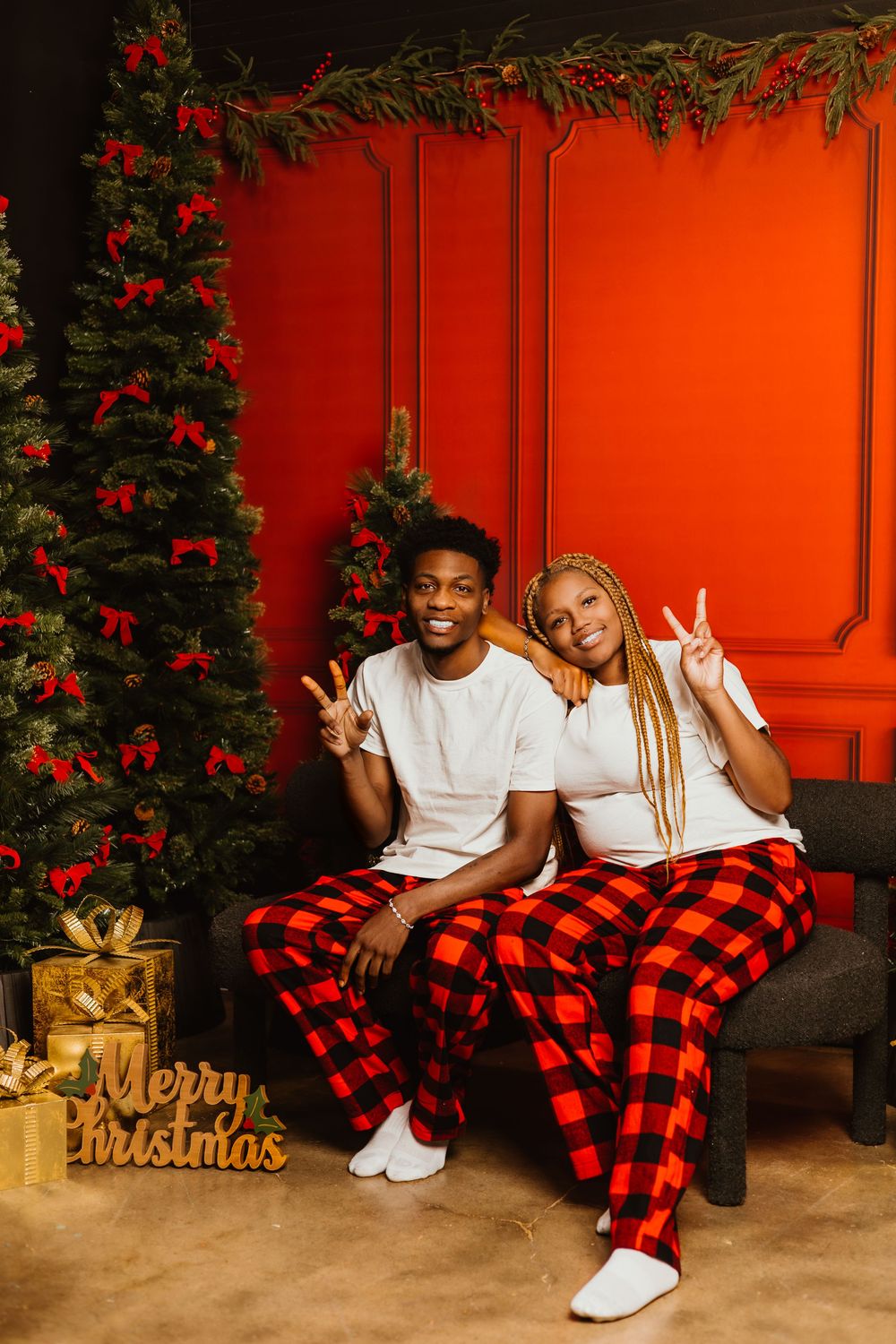 A festive Christmas photo setup with matching red and black plaid pajama pants against a bright red wall and holiday decor.