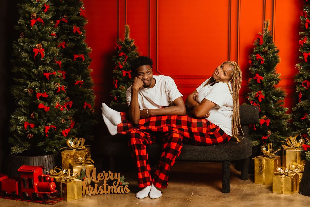A couple in buffalo plaid sits near a Christmas tree with gold presents and red decorations against a red wall.