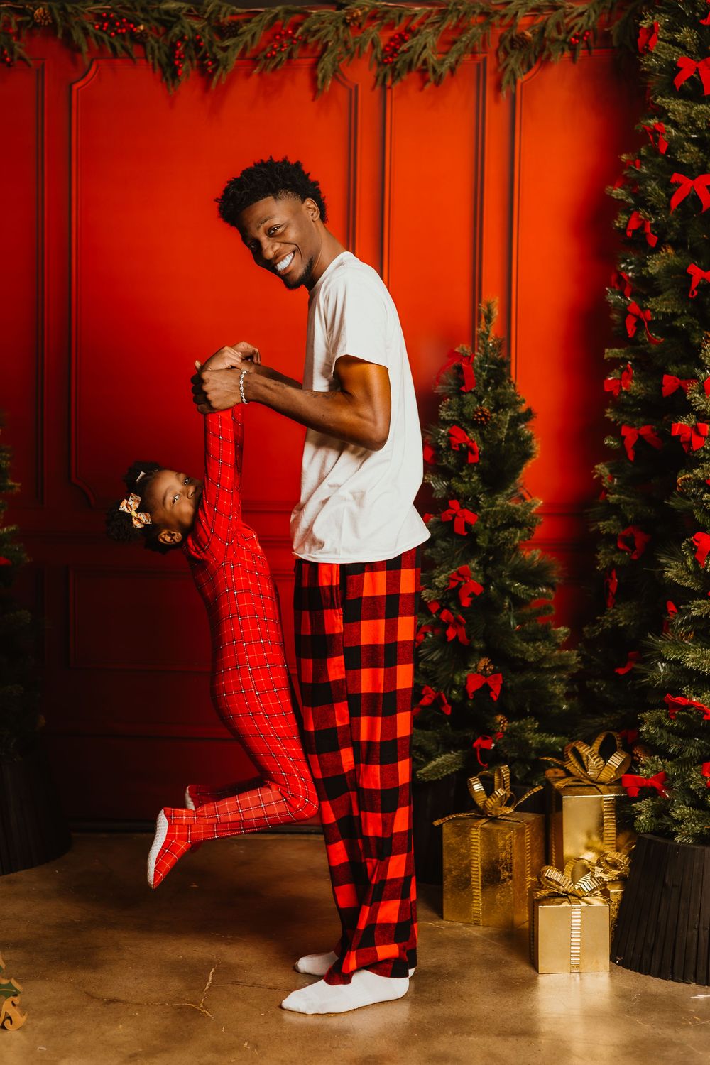Family members in matching buffalo plaid pajamas dance together in front of a Christmas tree and red wall.