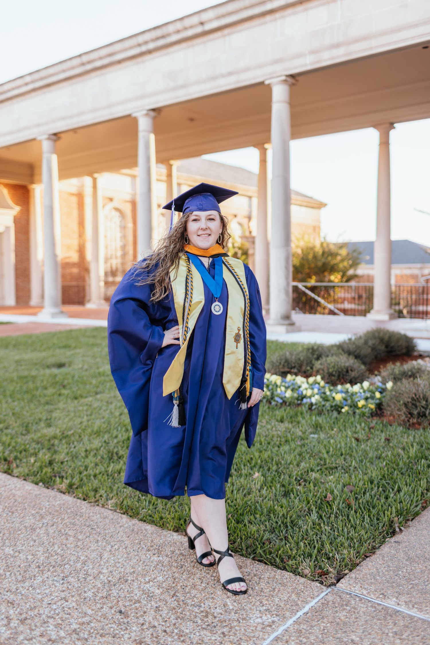 Graduate in blue cap and gown poses in front of columned building during golden hour sunset.