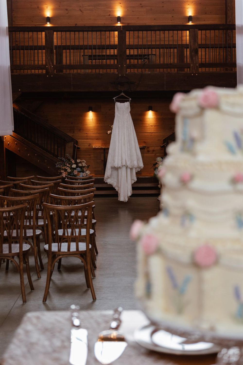 Wedding dress hangs in rustic venue with wooden chairs and tiered cake in foreground.