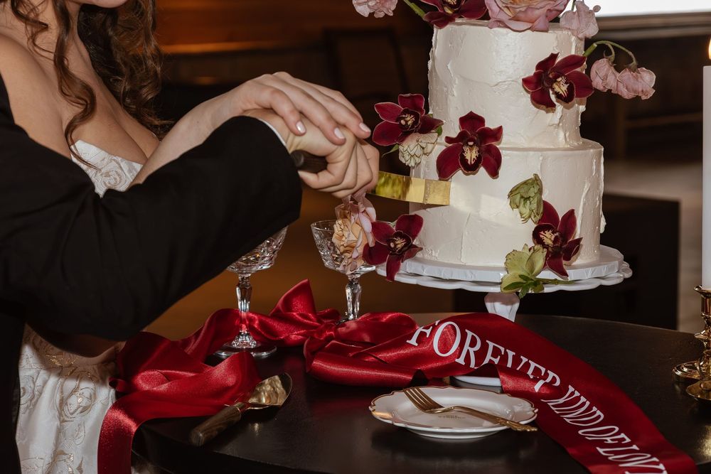 Wedding cake decorated with burgundy flowers being cut on dark table with red ribbon.