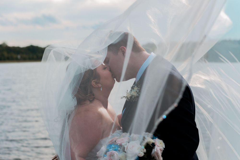 Bride and groom sharing a romantic kiss under a flowing wedding veil by a lakeside on their wedding day.