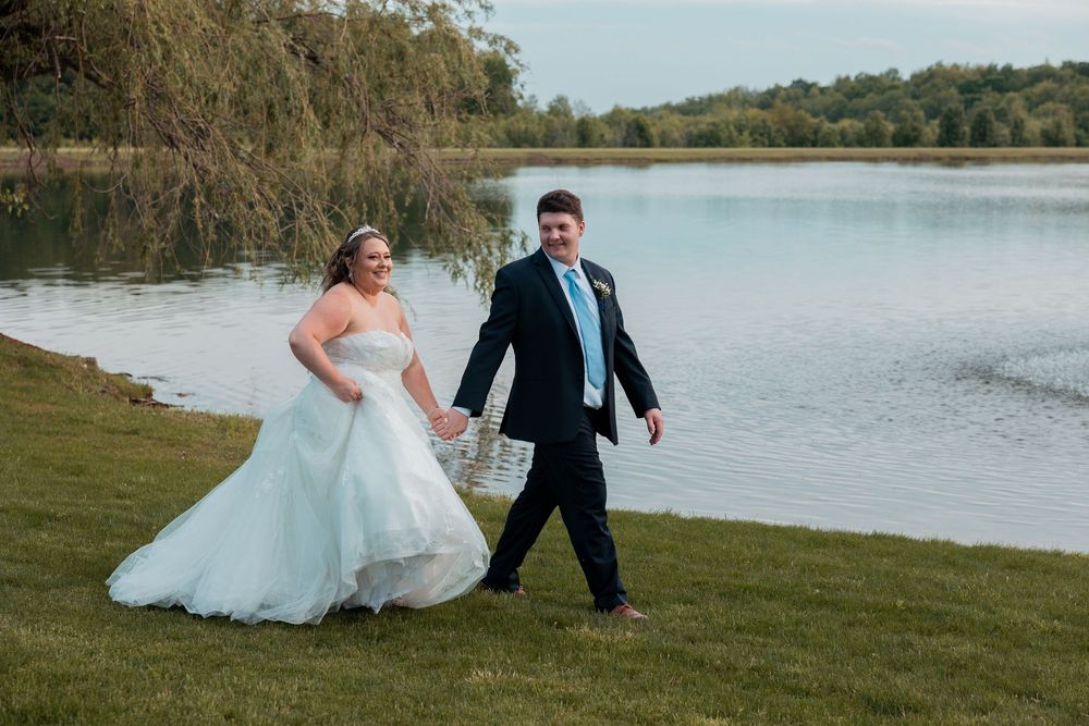 Bride in white gown and groom in navy suit hold hands walking by a serene lakeside on their wedding day.