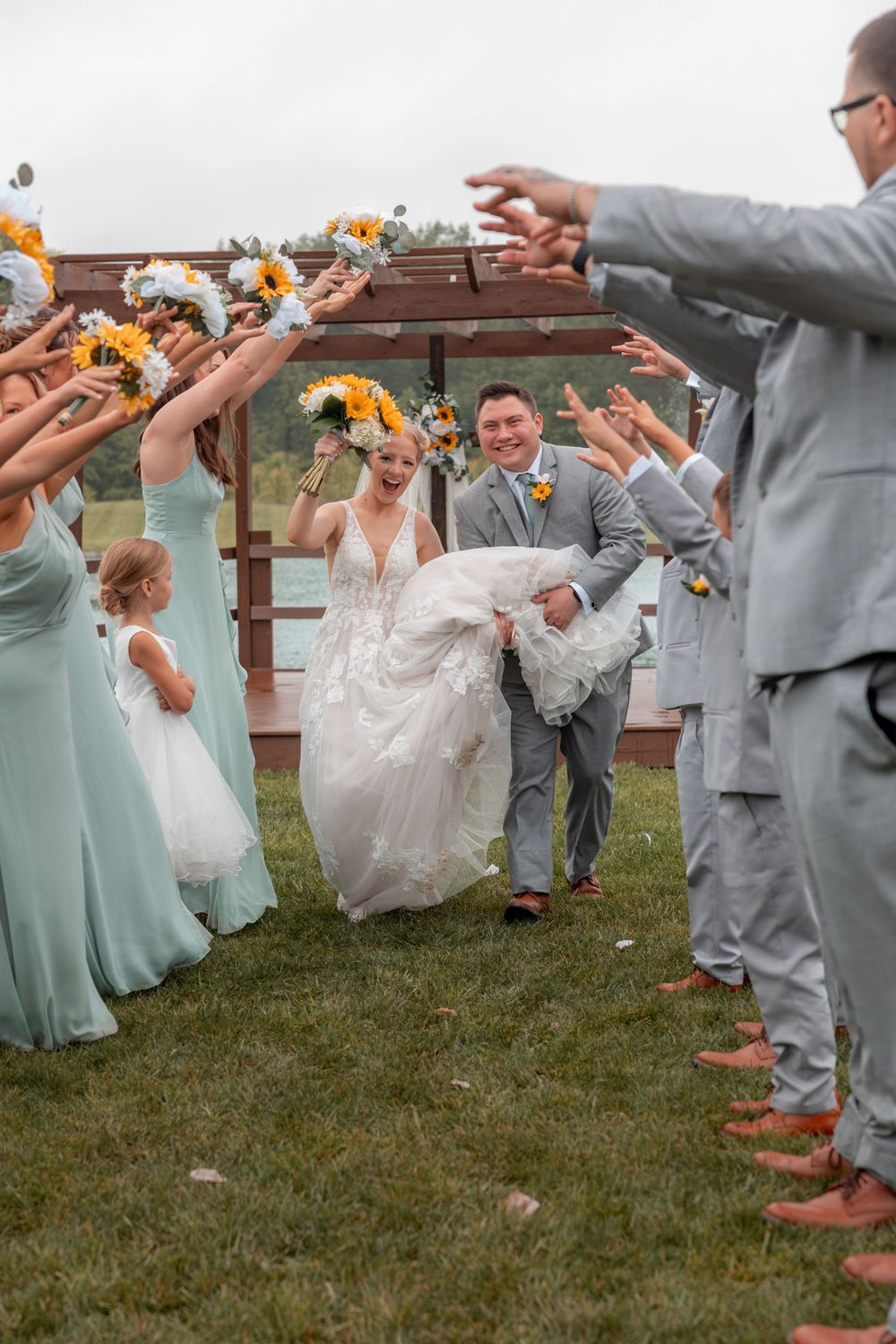 Joyful newlyweds exit outdoor wedding ceremony as bridesmaids in mint dresses and groomsmen in gray suits celebrate with raised bouquets.