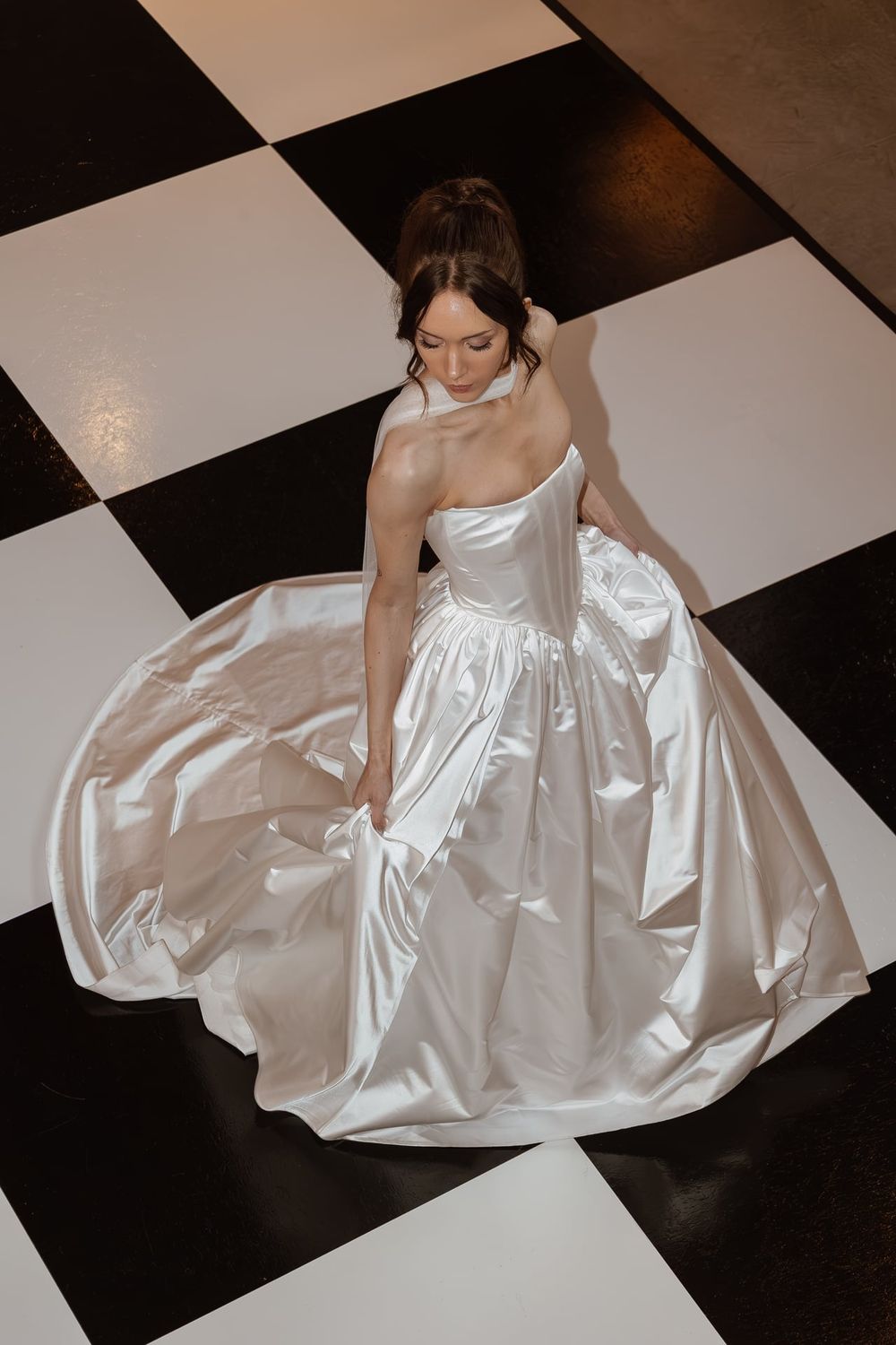Bride in white satin gown poses on black and white checkered floor.