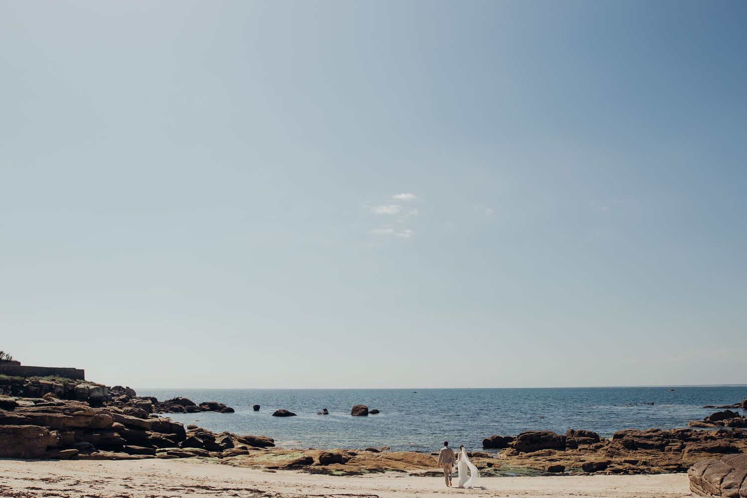 Two people stand on a rocky beach with calm blue ocean waters and clear sky in the background.