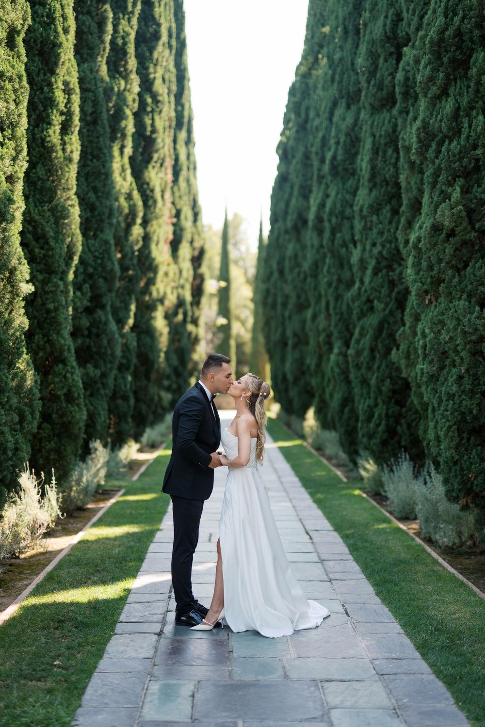 A romantic wedding couple shares intimate moments along a cypress tree-lined pathway in a formal garden setting.