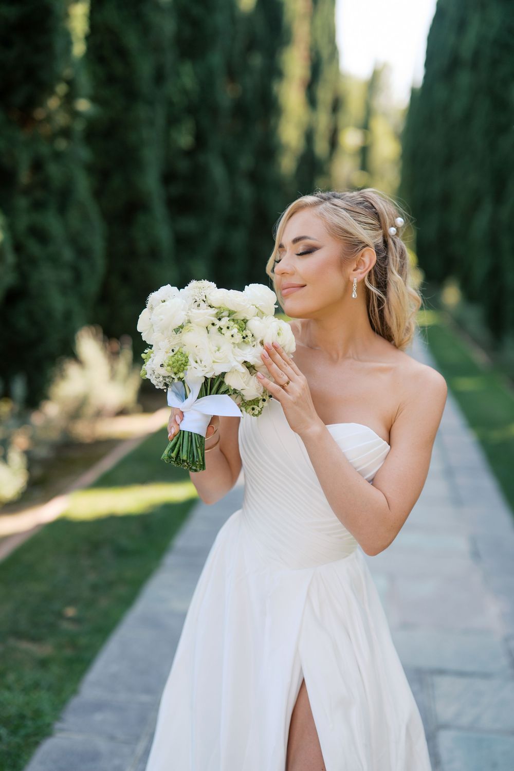 Bride in flowing white gown poses with her bouquet of white flowers along a cypress tree-lined path.