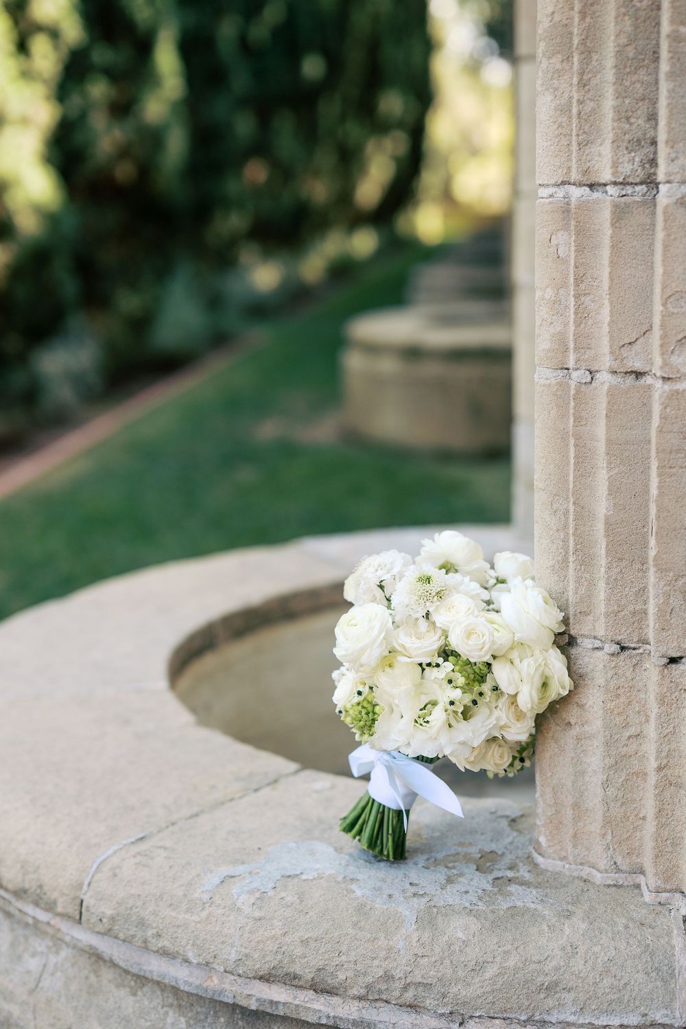 A delicate white bridal bouquet with roses and baby's breath tied with a light blue ribbon rests on a stone ledge.