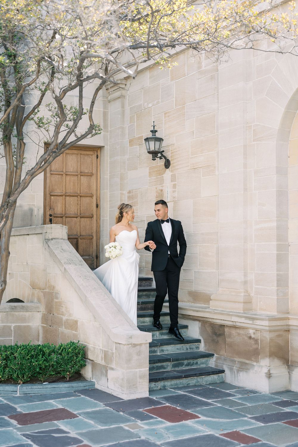 A couple in formal wedding attire stands on elegant stone steps outside a church with white walls and a lantern.