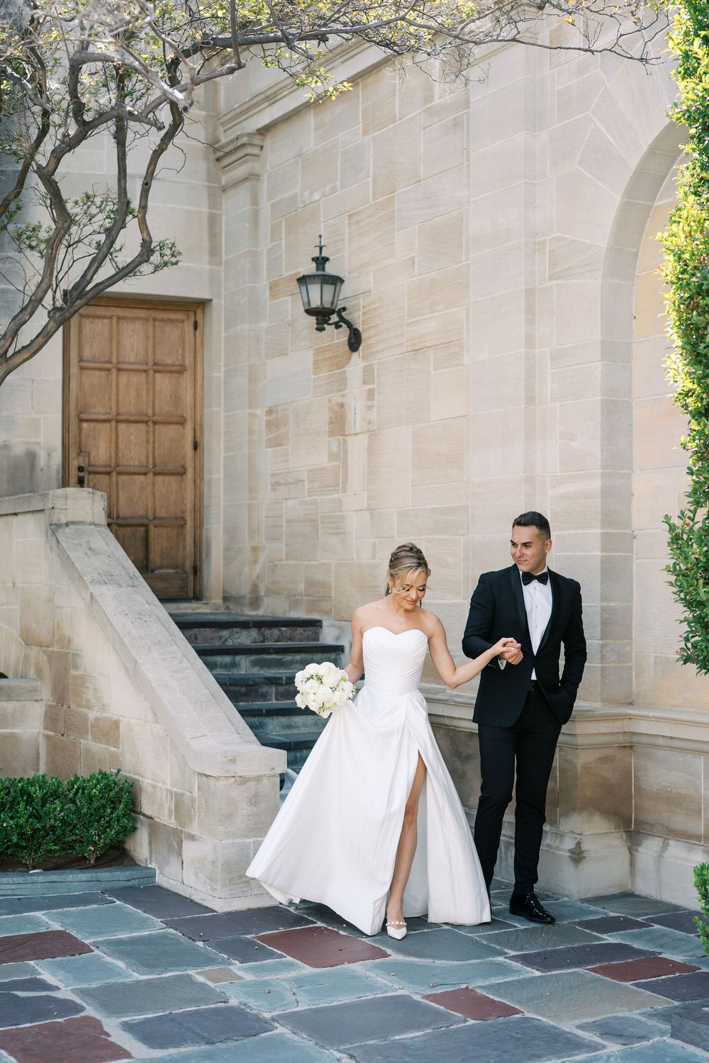 Elegant wedding couple stands in front of stone archway with wooden door and vintage lantern at luxury venue.