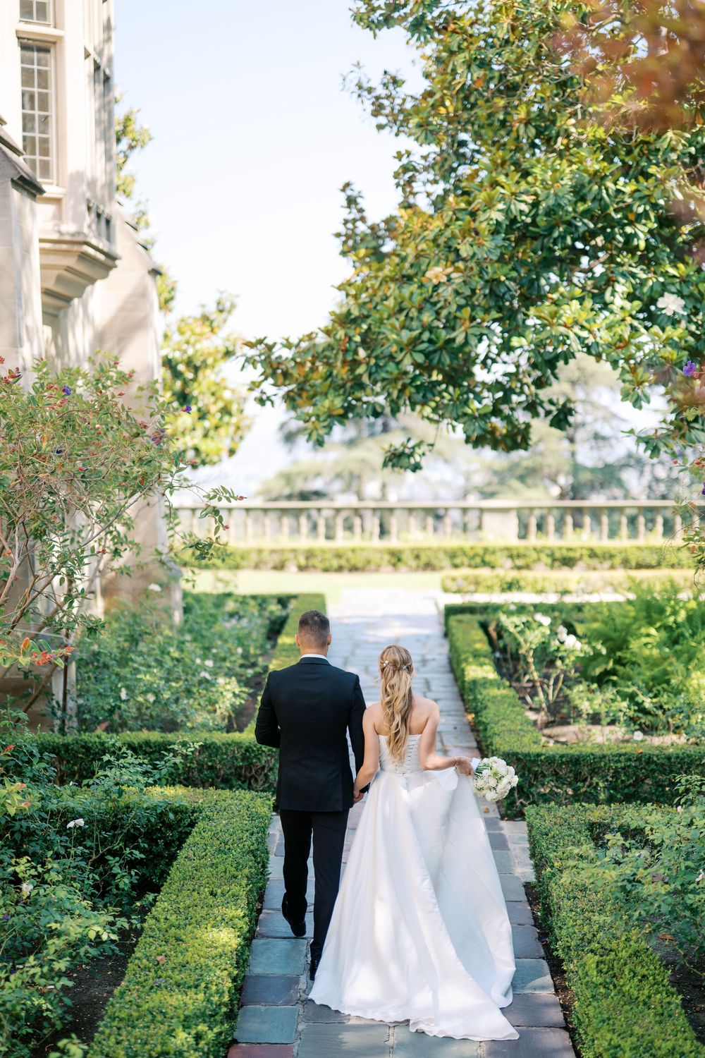 A couple walks down a garden path surrounded by manicured hedges and greenery at an elegant estate venue.