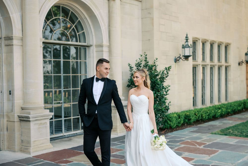 Elegant wedding couple poses in front of grand arched windows and stone columns at a historic venue.