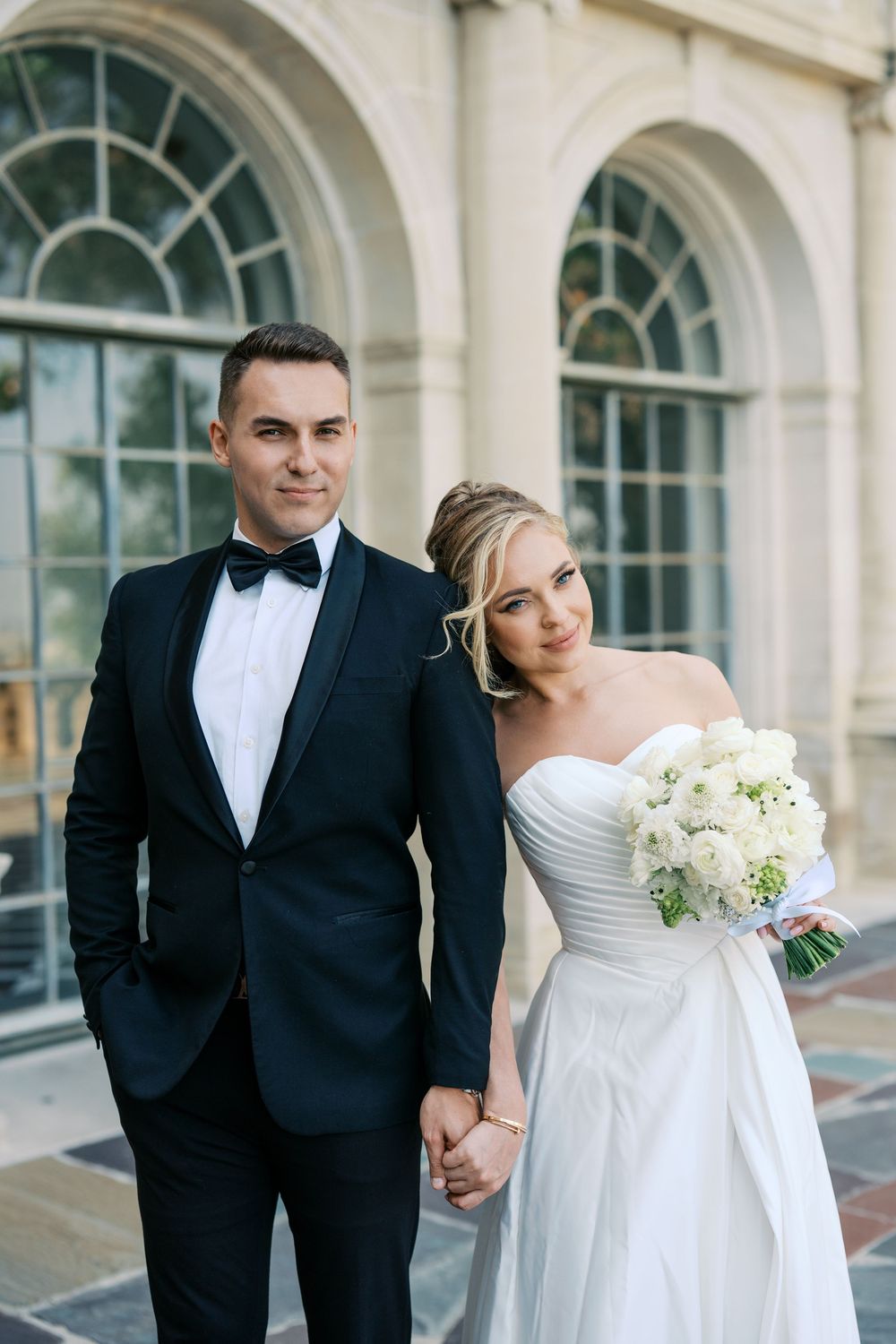 A bride and groom pose together in formal wedding attire in front of elegant arched windows at a historic venue.