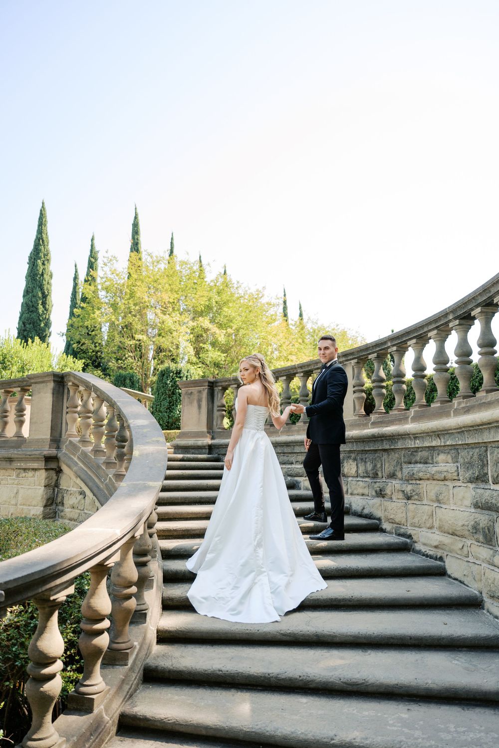 A couple stands on elegant stone steps surrounded by cypress trees and classical architecture.