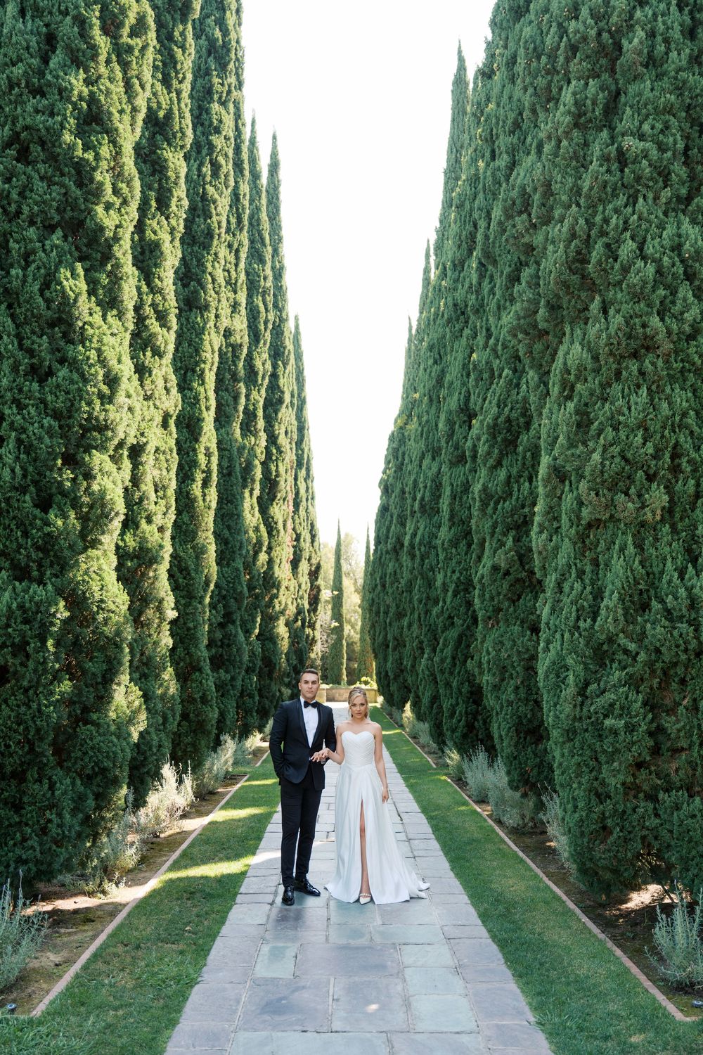 A romantic garden pathway lined with tall cypress trees and stone pavers creates an elegant outdoor setting.