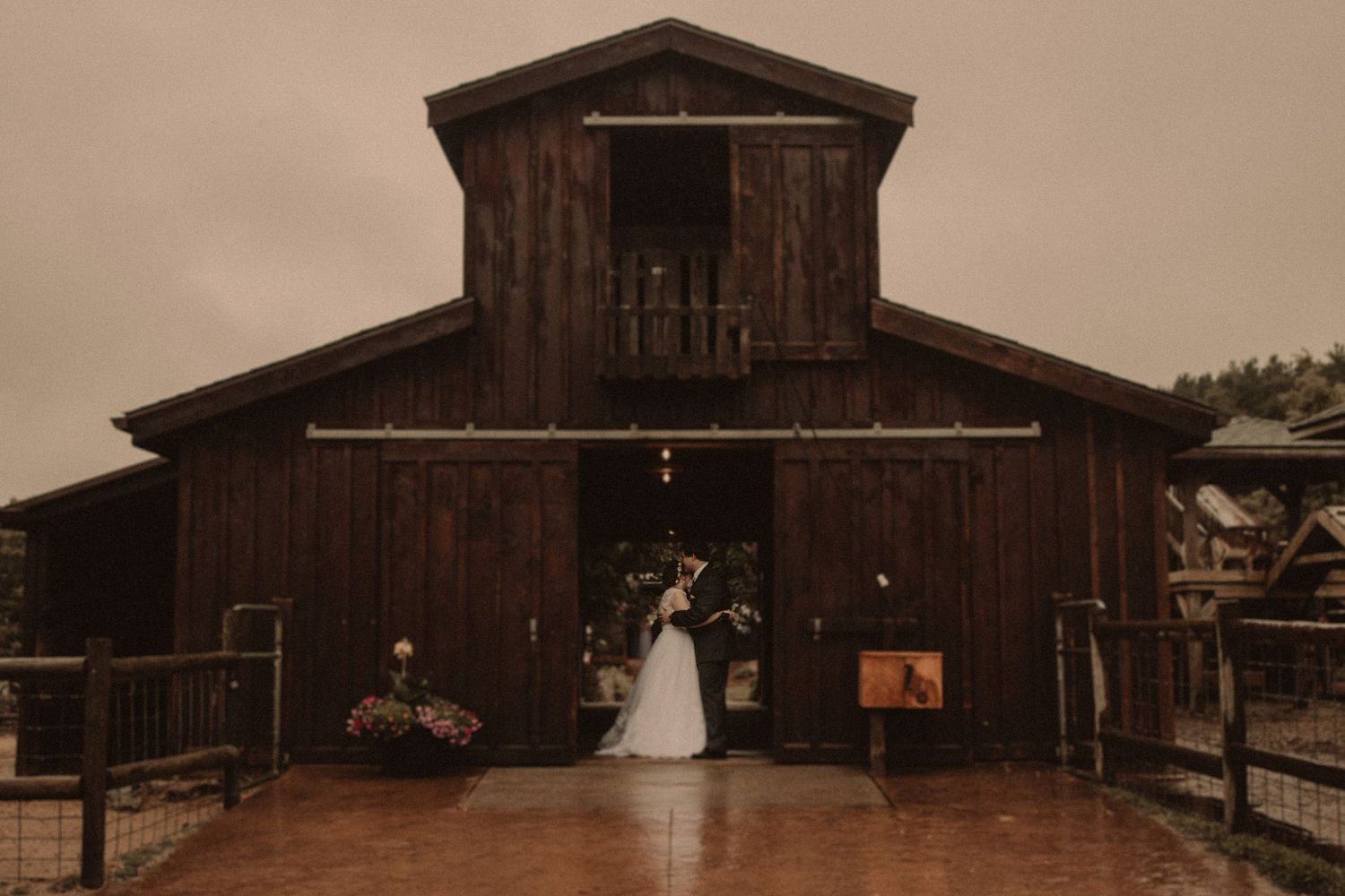 A rustic brown wooden barn with weathered siding stands against a sepia-toned rural landscape.