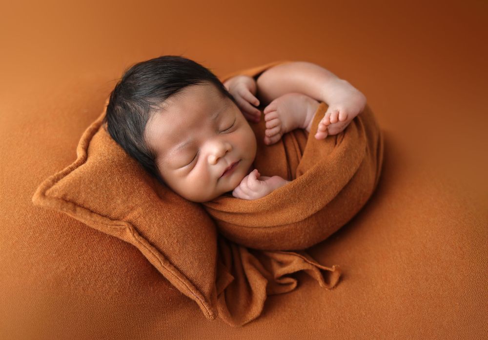 Sleeping newborn baby wrapped in soft brown fabric on warm terracotta background in artistic photo series.
